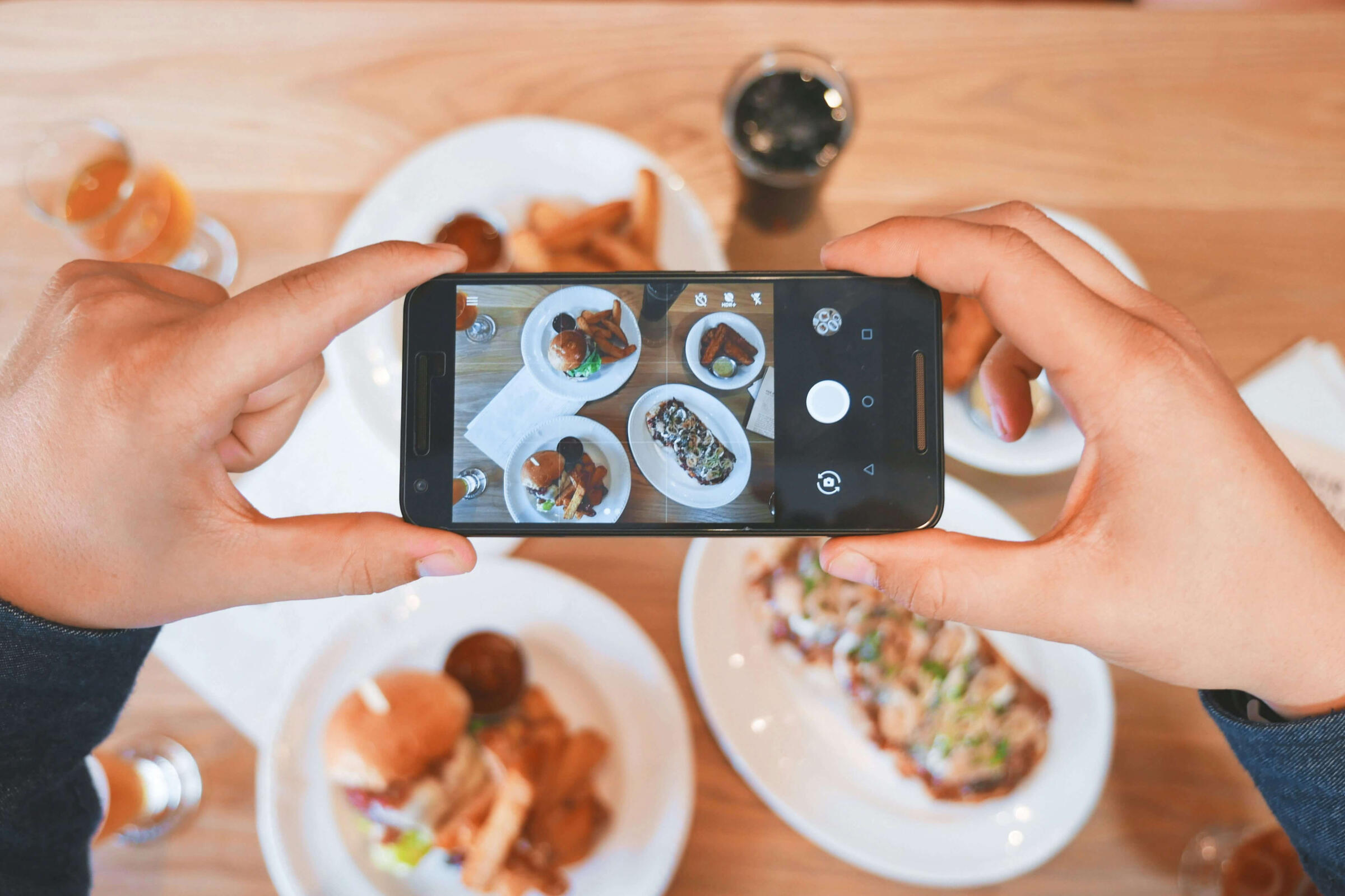 Bistrón social media & strategy service Hands holding a smartphone taking an overhead photo of multiple plates of food on a wooden table.